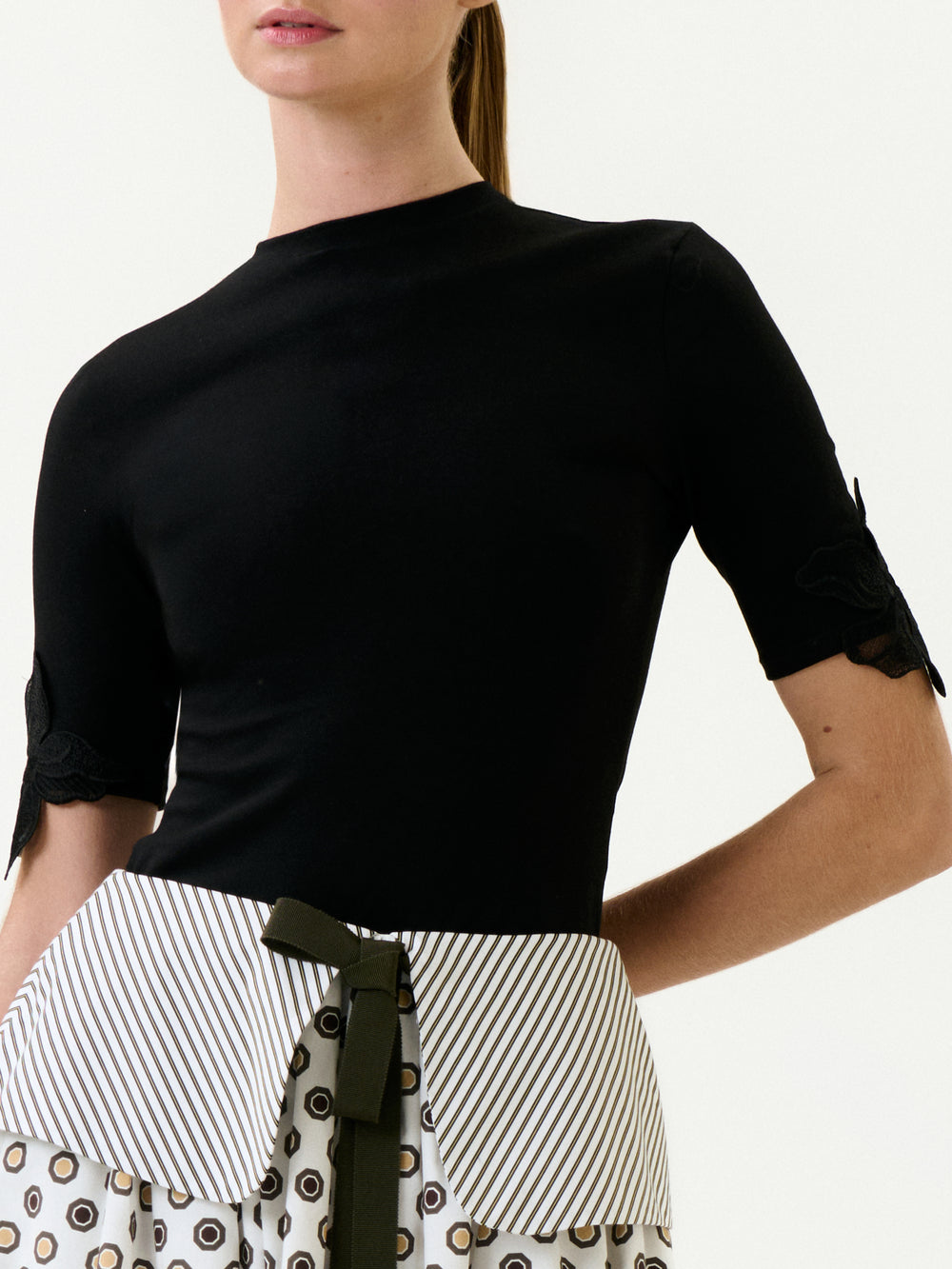 A woman wears the Walda T-Shirt, a fitted black short-sleeve top, paired with a high-waisted skirt featuring Italian fabric, vertical stripes, circular patterns, and a dark bow. Her hair is pulled back; her face is partially out of frame.