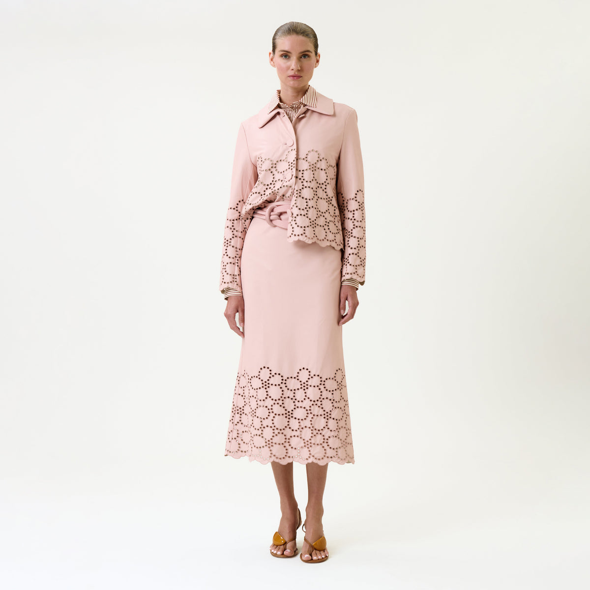 A woman stands against a white background wearing the Atira Skirt and a matching light pink shirt with laser-cut floral patterns, paired with yellow heeled sandals.