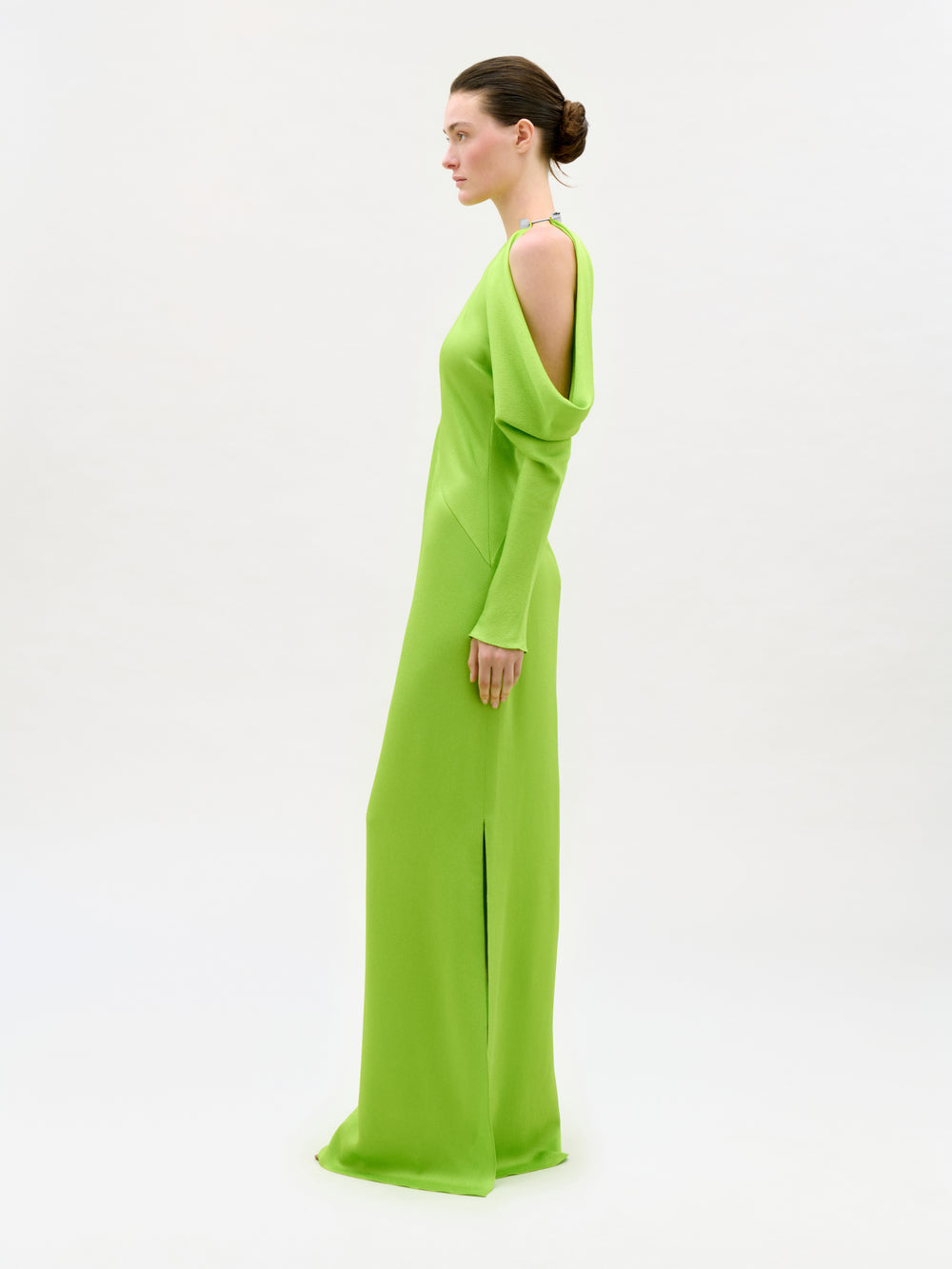 A woman with brown hair in a low bun stands in profile against a plain white background, wearing the bright green Antonia Dress—a column silhouette maxi with open shoulders and long sleeves.