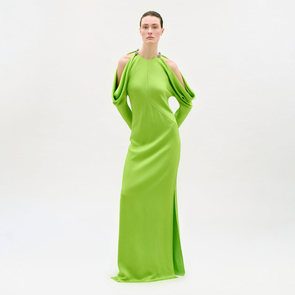 A person poses before a white backdrop in the Antonia Dress, a bright green Italian maxi with dramatic draped sleeves, shoulder cutouts, high neckline, and a straight column silhouette.