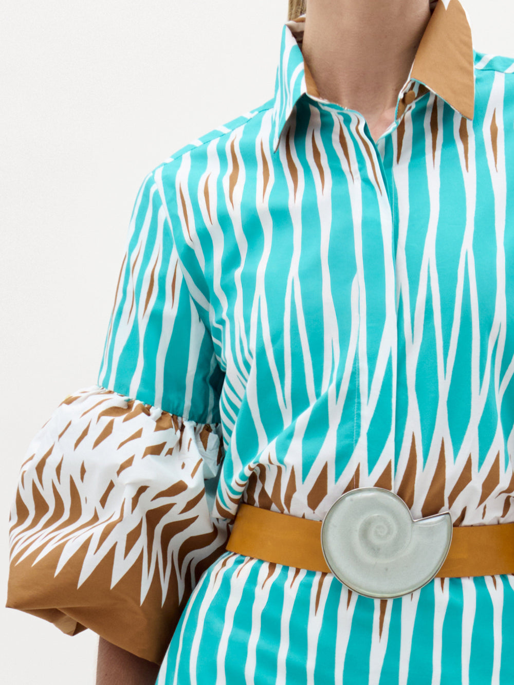 A woman stands against a plain white background wearing the Helina Blouse—a turquoise and white patterned top made from Italian fabric with brown details, puffy short sleeves, and a beige belt, accessorized with clear bracelets.