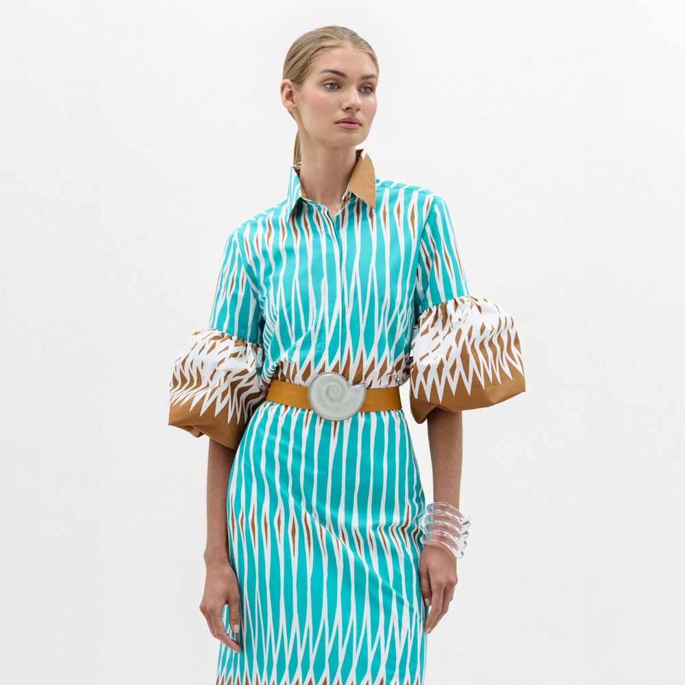 A woman stands against a plain white background wearing the Helina Blouse—a turquoise and white patterned top made from Italian fabric with brown details, puffy short sleeves, and a beige belt, accessorized with clear bracelets.
