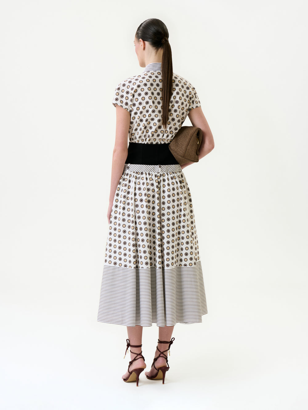 A woman with a low ponytail and strappy heels stands facing away on a plain white background, wearing the Odalis Dress—a patterned 100% cotton midi with a black waistband—and holding a textured brown clutch.