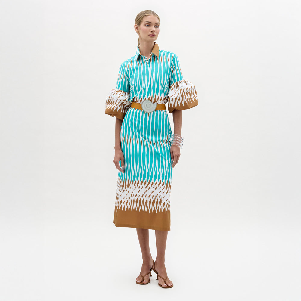 A woman stands against a plain white background wearing the Atira Skirt, a high-waisted, turquoise, white, and brown patterned midi skirt styled with puffed sleeves, a wide belt, strappy sandals, and bracelets.