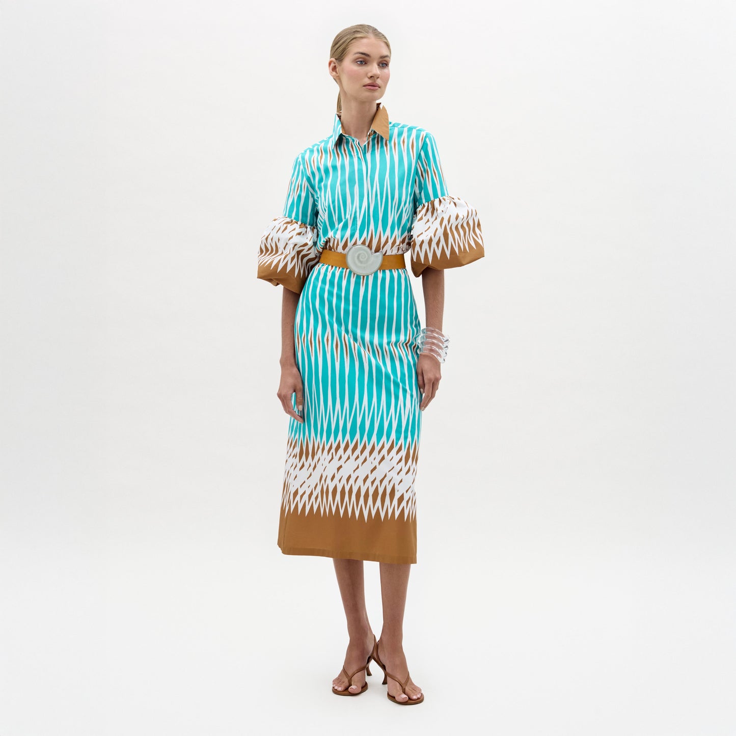 A woman stands against a plain white background wearing the Atira Skirt, a high-waisted, turquoise, white, and brown patterned midi skirt styled with puffed sleeves, a wide belt, strappy sandals, and bracelets.
