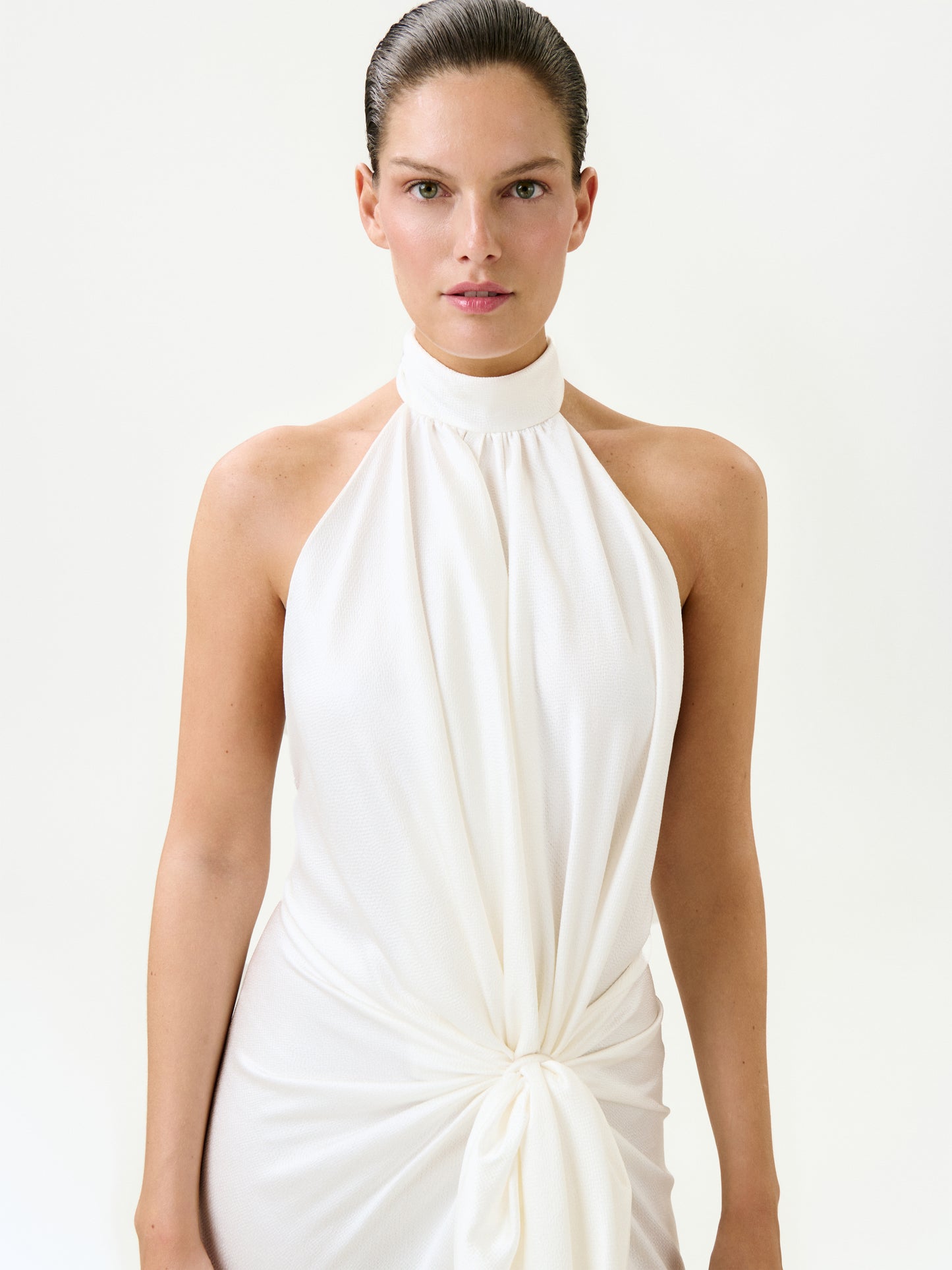 A woman with slicked-back hair models the Marisela Dress—a sleeveless white halter-neck maxi made in Portugal with a knotted waist detail—standing against a plain light background.