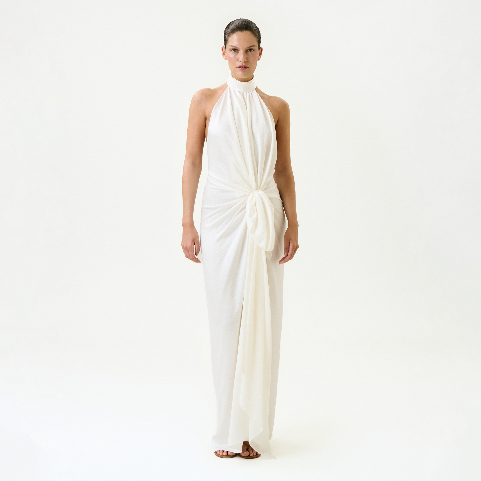 A woman stands facing forward in the Marisela Dress—a sleeveless, floor-length white maxi with a high neckline and draped knot waist, expertly crafted from fine Italian fabric—against a plain white background.