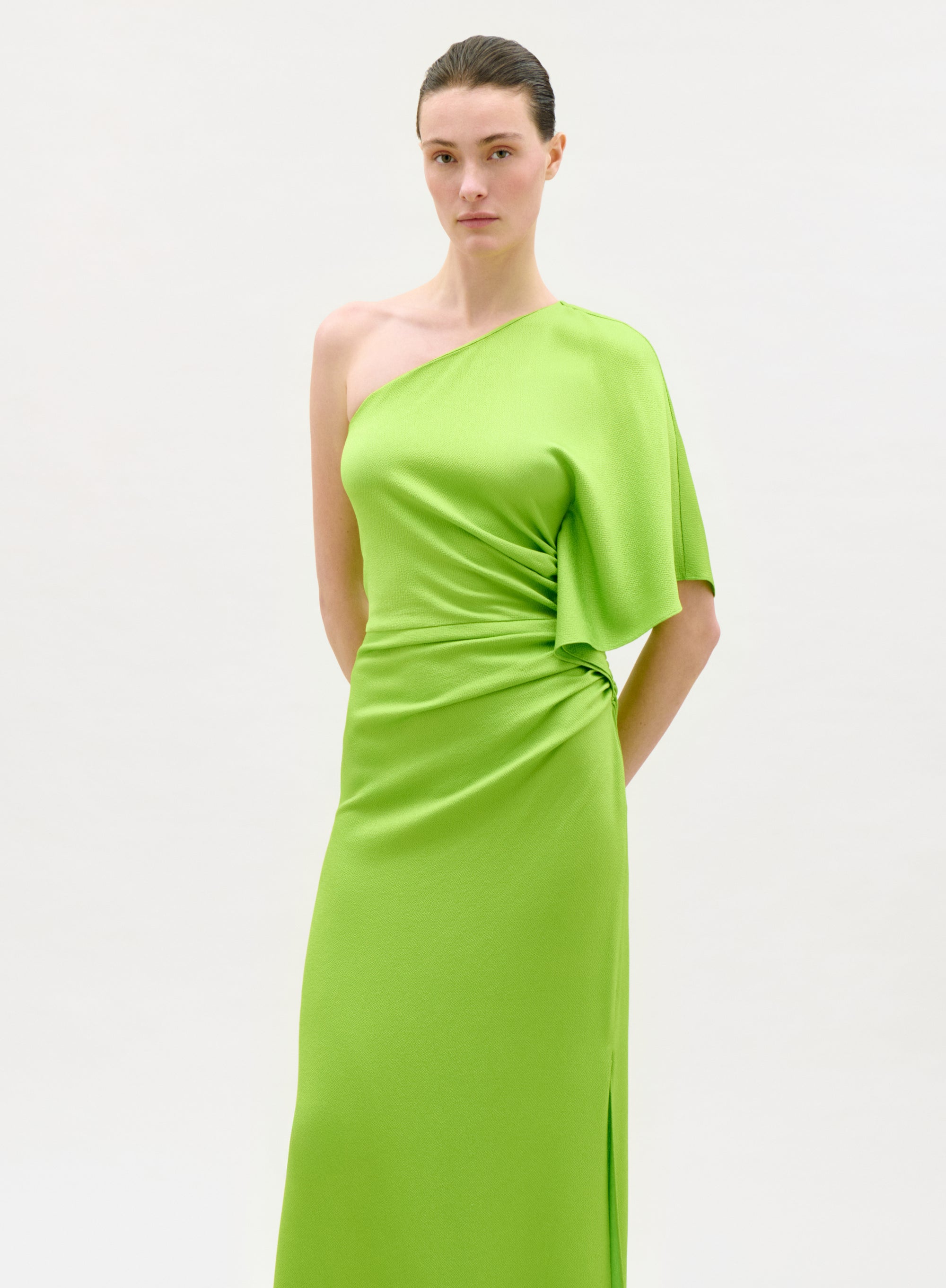 A woman models the Doli Dress, a vivid green one-shoulder maxi crafted from Italian fabric with a draped asymmetric sleeve and gathered waist detail, set against a plain light background.