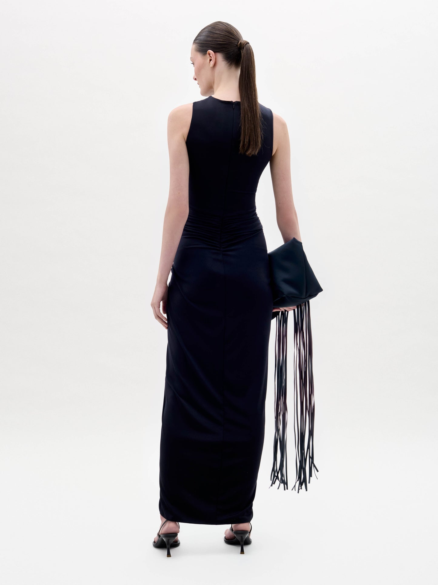 Woman wears the Leni Dress, a sleeveless black dress with a high slit, and holds a large black fringed clutch, standing against a plain white background.