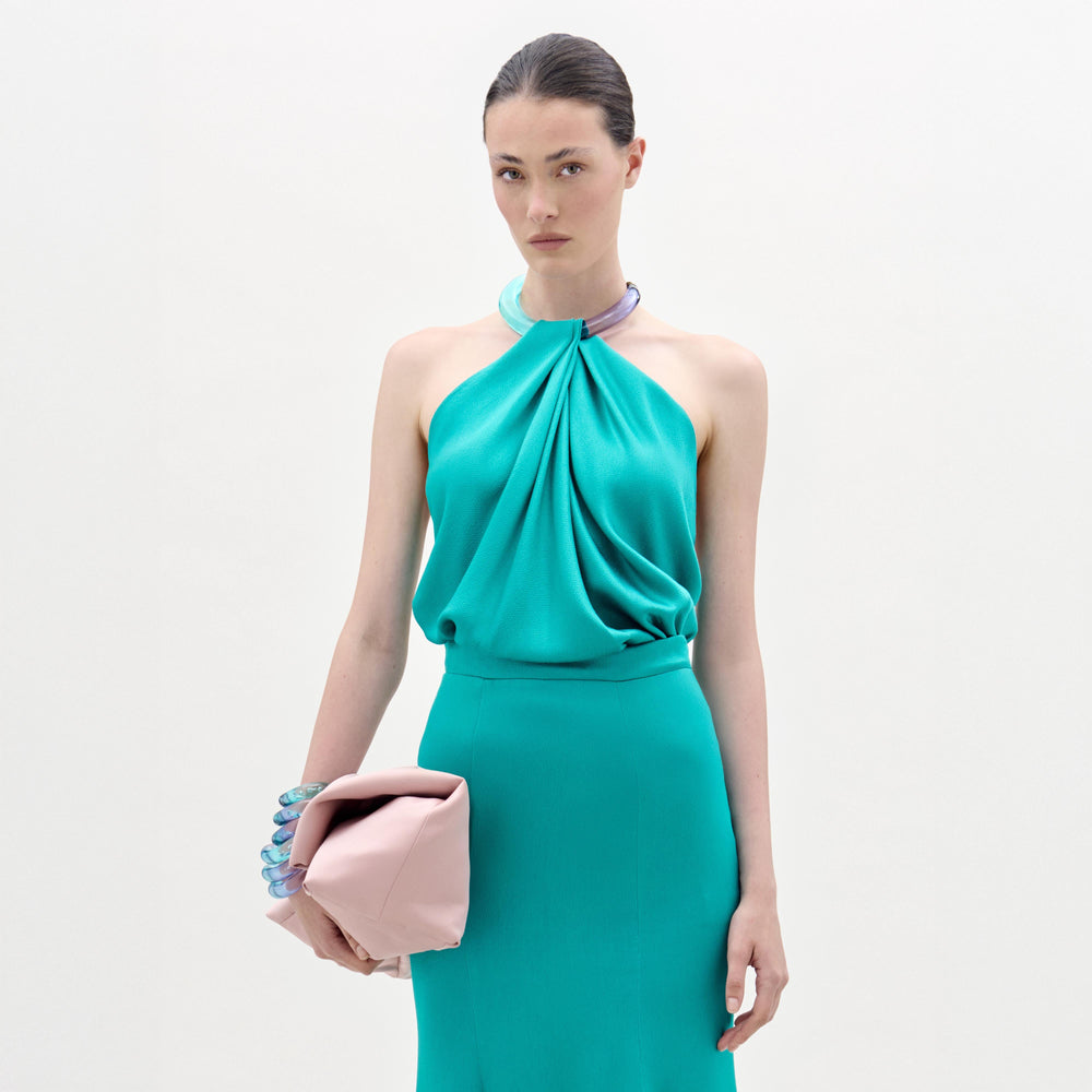 A woman in the Dianela Blouse, made from luxurious Italian fabric in teal, holds a pale pink clutch against a plain white background.