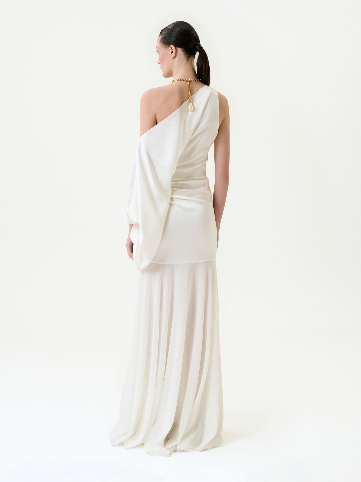 A woman with her dark hair in a ponytail stands against a plain white background, wearing the Emilia Dress—a flowing off-shoulder silk maxi with a single sleeve—and a gold necklace.