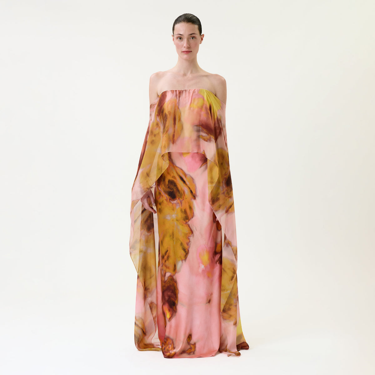A person stands against a plain background wearing the strapless, flowing Lovane Dress, featuring a pink, yellow, and brown abstract pattern.