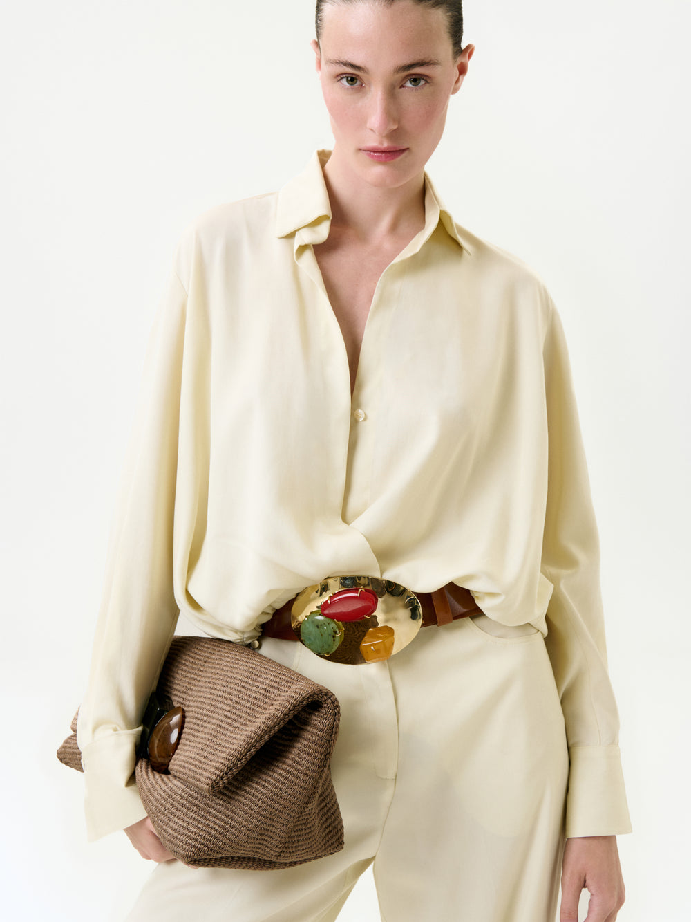 A woman in the loose, cream-colored Saula Blouse and matching Italian-fabric pants holds a textured brown clutch. She wears a bold, colorful statement belt buckle and stands against a plain white background.