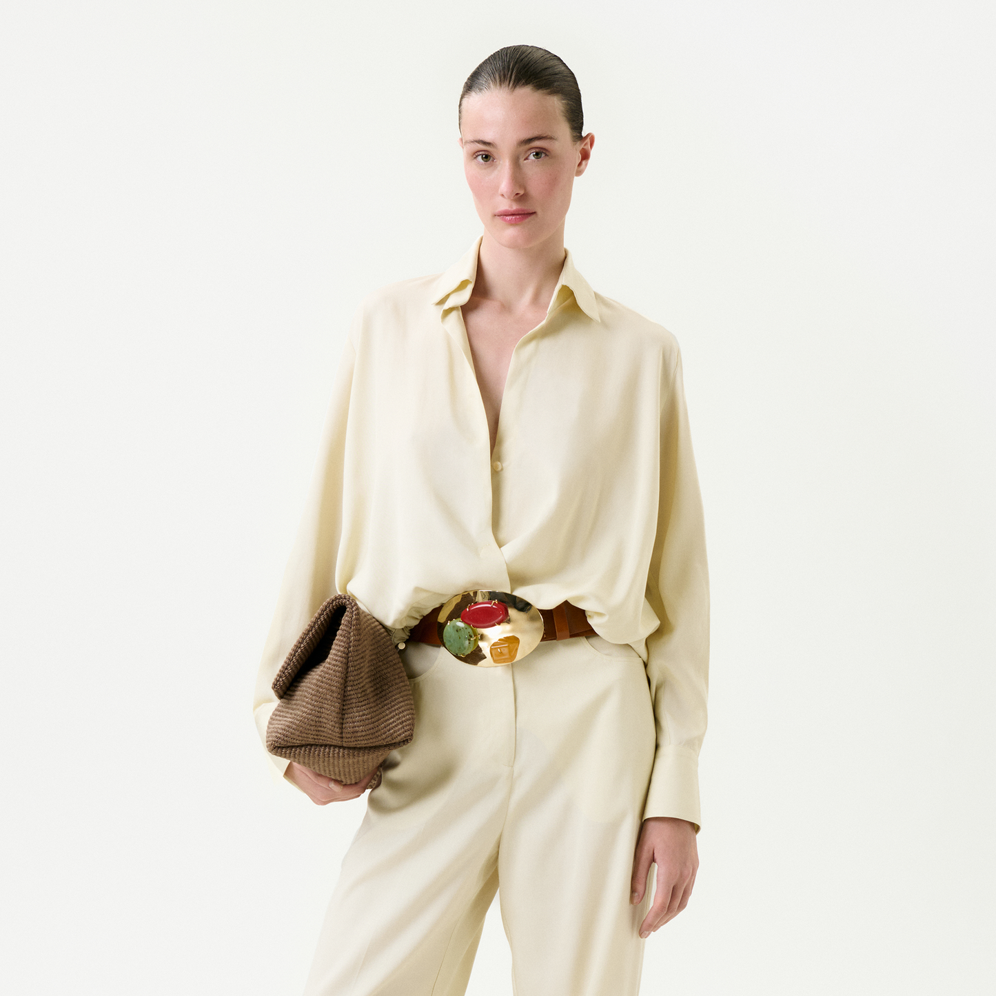 A woman with slicked-back hair wears the Saula Blouse, a loose, cream piece crafted from premium Italian fabric. She pairs it with matching trousers, a wide belt with a colorful buckle, and holds a textured clutch against a white background.