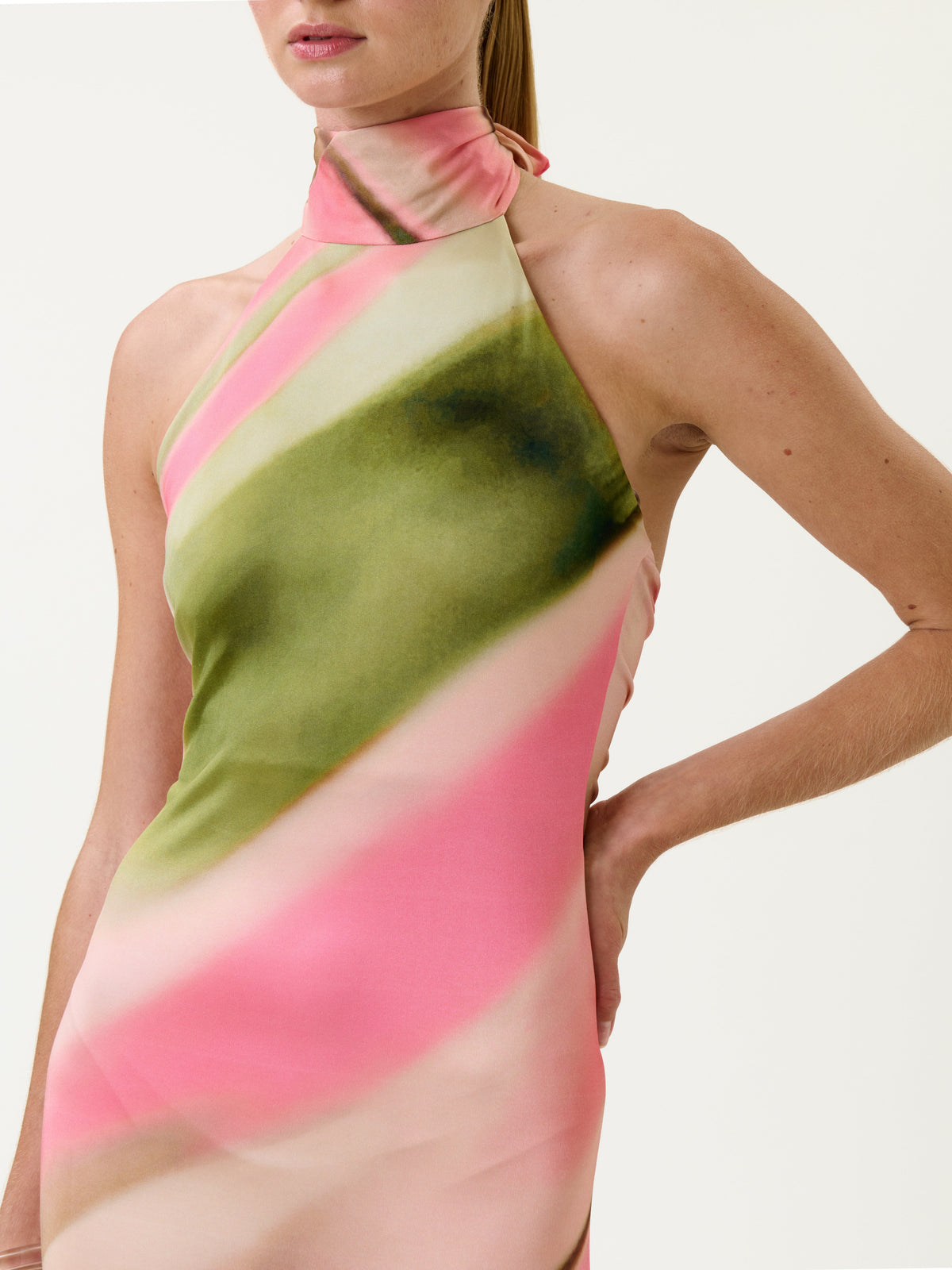 A person wearing the Maika Dress—a sleeveless halter-neck style featuring pink, green, and cream diagonal patterns—stands with one hand on their hip against a plain background.