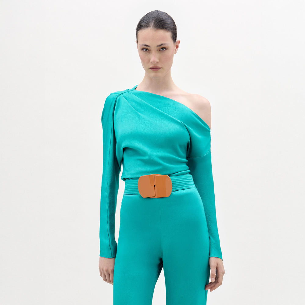 A woman wears the Viola Blouse, a teal one-shoulder top with draped detailing and an orange belt, standing against a plain white background.