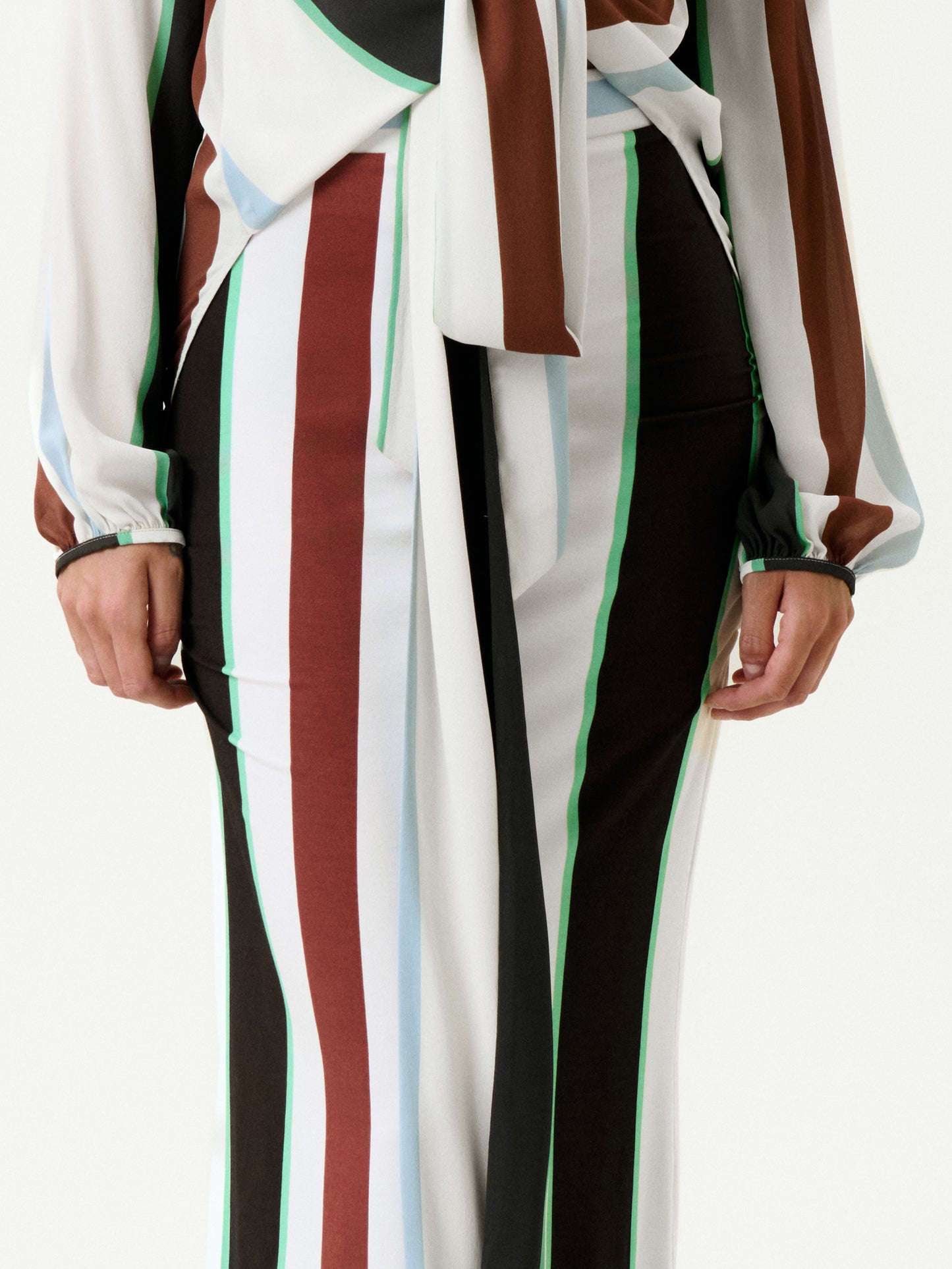 A person wears the Toni Skirt—a flowing, draped skirt with vertical stripes in brown, green, white, light blue, and black. Made in Colombia from Italian fabric, it's styled here with a matching long-sleeve top. Hands are relaxed at the sides.