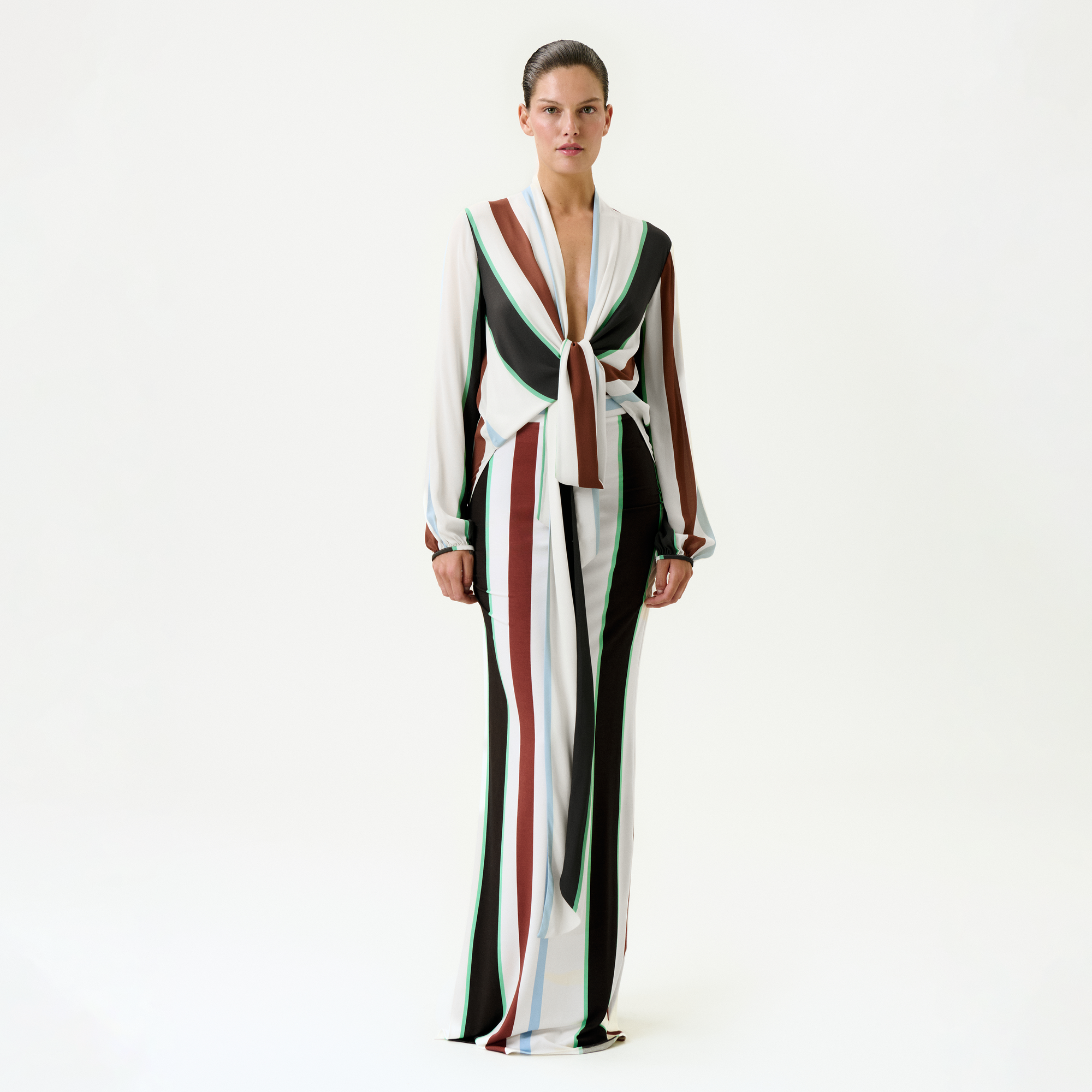 A woman stands against a plain white background wearing the Toni Skirt, a slim fit, floor-length skirt with bold vertical stripes in brown, green, white, and black, featuring a front knot detail at the waist.