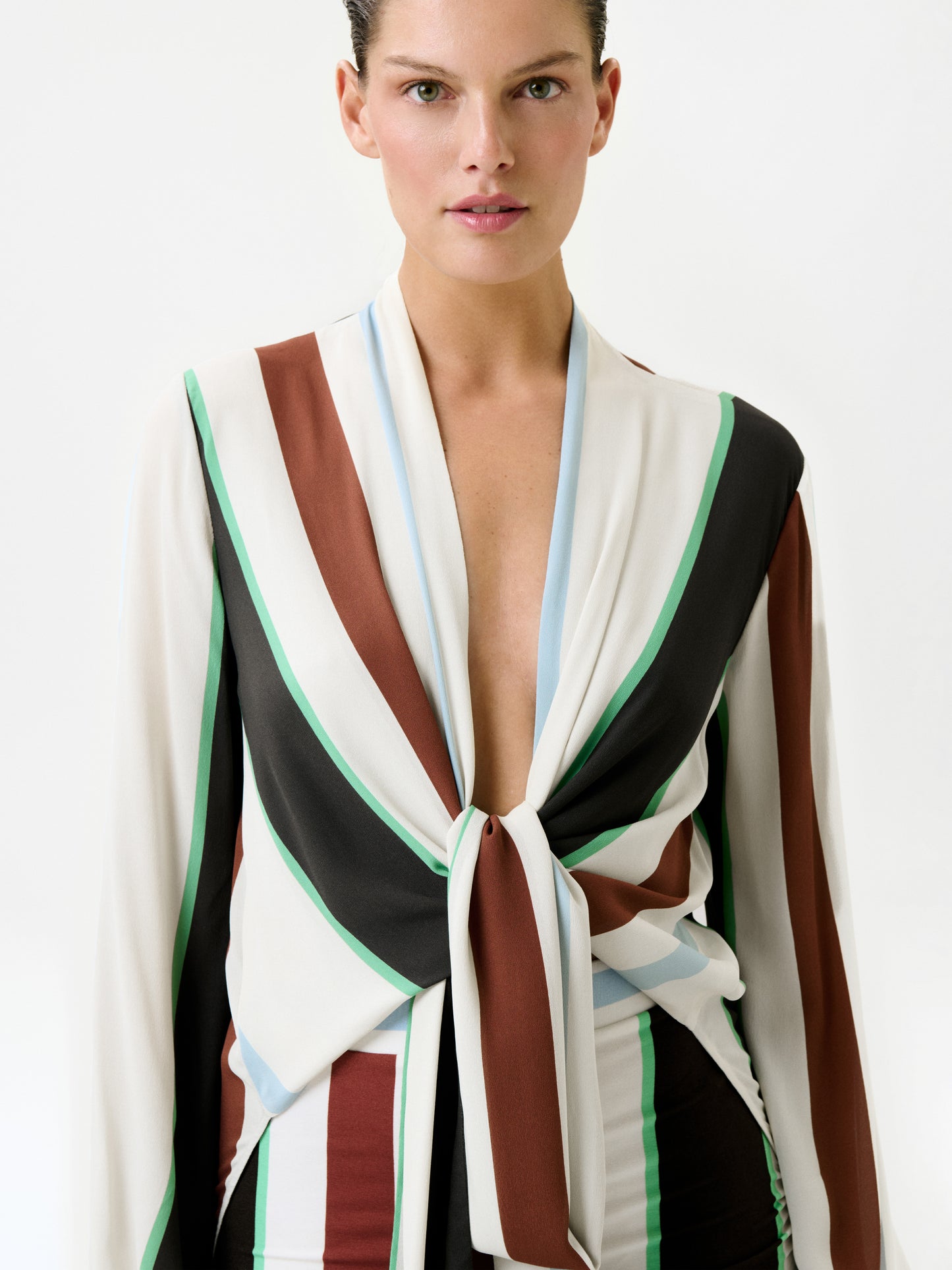 A person wearing the Honey Blouse, a relaxed fit long-sleeve top featuring vertical stripes in brown, white, black, green, and light blue, stands facing forward against a plain white background. Made from 100% viscose Italian fabric, the blouse is tied at the front and reveals part of the chest.