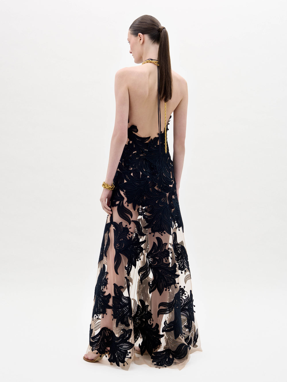 A woman wearing the Mab Dress, a black sleeveless gown with sheer nude panels and black floral patterns, accessorized with gold necklaces and bracelets, stands against a plain white background.
