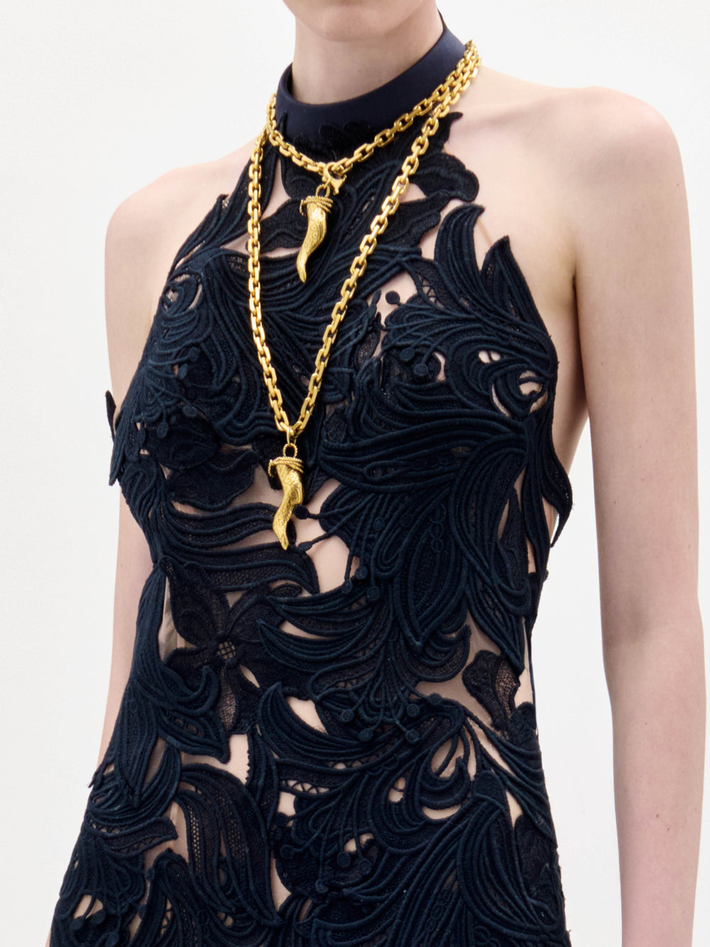 A woman wearing the Mab Dress, a black sleeveless gown with sheer nude panels and black floral patterns, accessorized with gold necklaces and bracelets, stands against a plain white background.