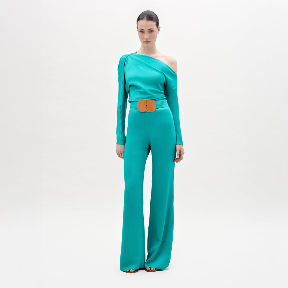 A woman stands against a plain background wearing the Palermo Pant, a teal Italian-fabric one-shoulder jumpsuit with wide-leg pants and a large tan belt.