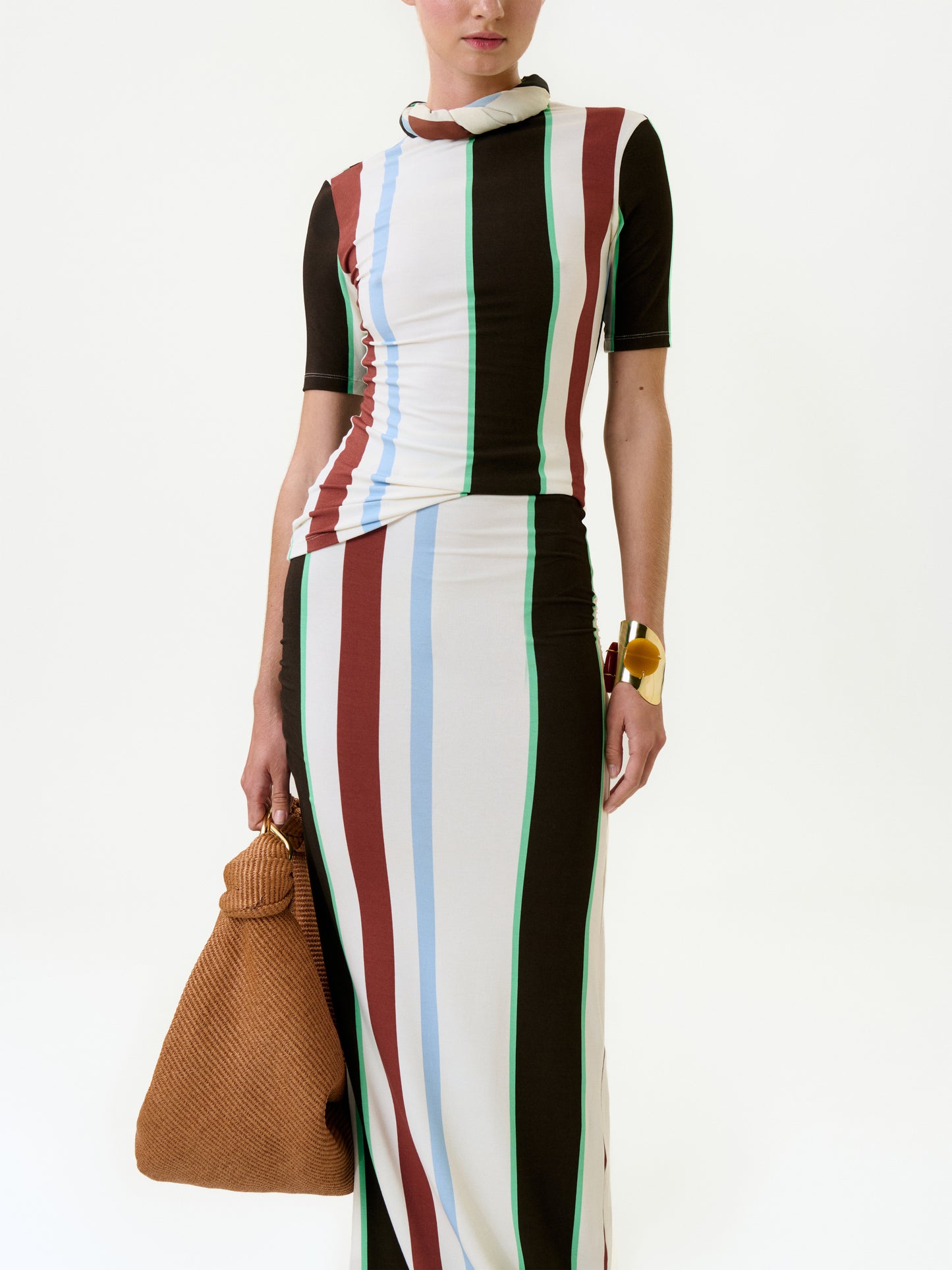 A person wears the Rowena Top, a short-sleeve piece made from Italian fabric with vertical brown, blue, green, and white stripes, styled with a large textured brown bag and chunky gold bracelet against a plain white background.