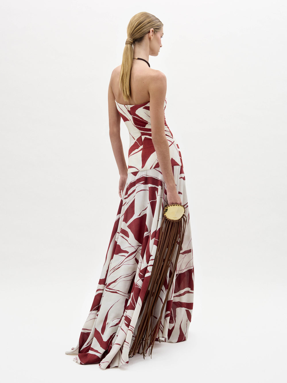 A woman stands wearing the Muriel Dress, a strapless, floor-length gown with a red and white abstract pattern, holding a brown fringe accessory.