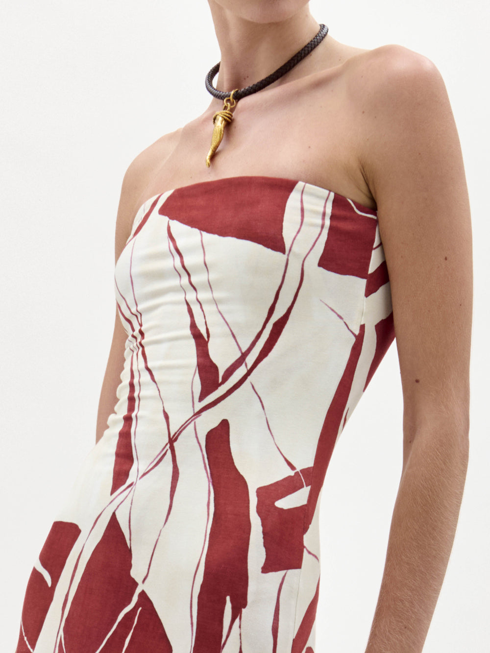 A woman stands wearing the Muriel Dress, a strapless, floor-length gown with a red and white abstract pattern, holding a brown fringe accessory.