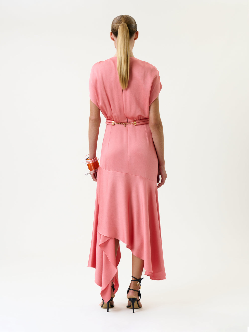 A woman stands facing away, wearing the Protea Dress—a pink, asymmetrical style paired with a gold belt and dark heels. Her hair is styled in a ponytail, and she wears several bracelets on her right wrist. The background is plain white.