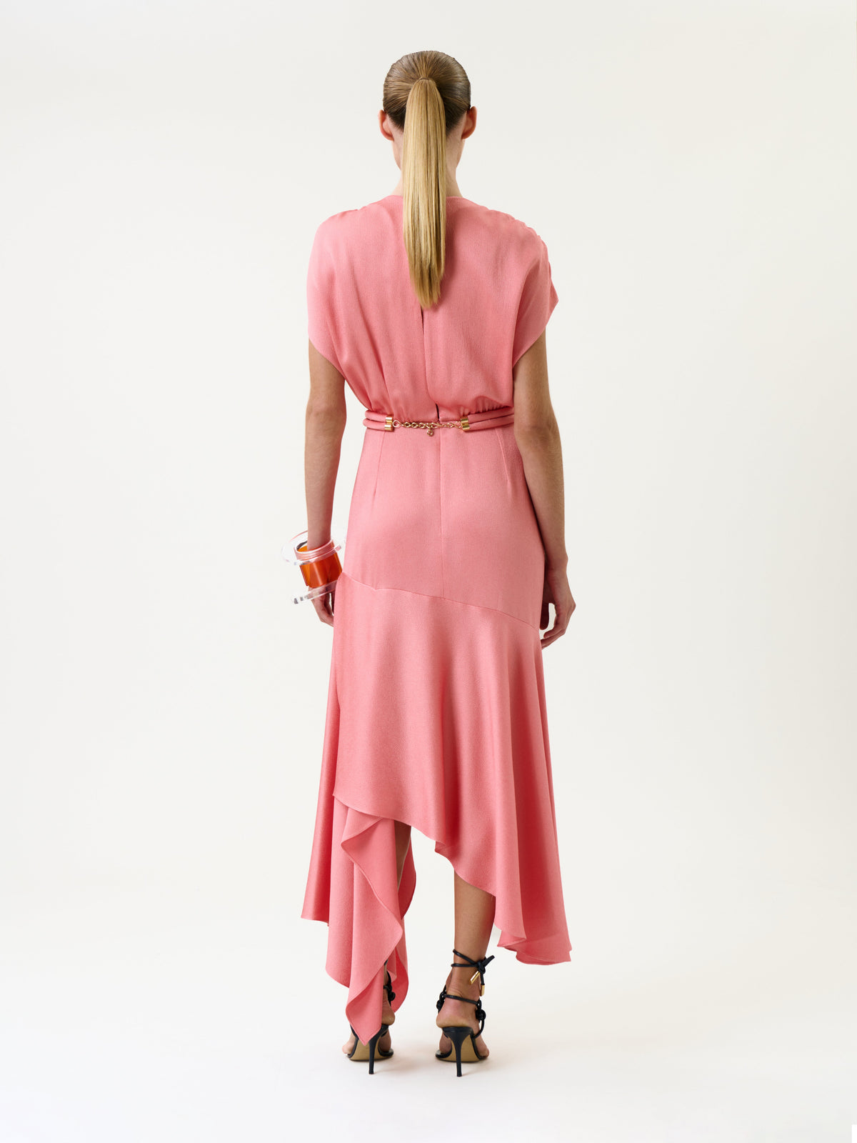 A woman stands facing away, wearing the Protea Dress—a pink, asymmetrical style paired with a gold belt and dark heels. Her hair is styled in a ponytail, and she wears several bracelets on her right wrist. The background is plain white.