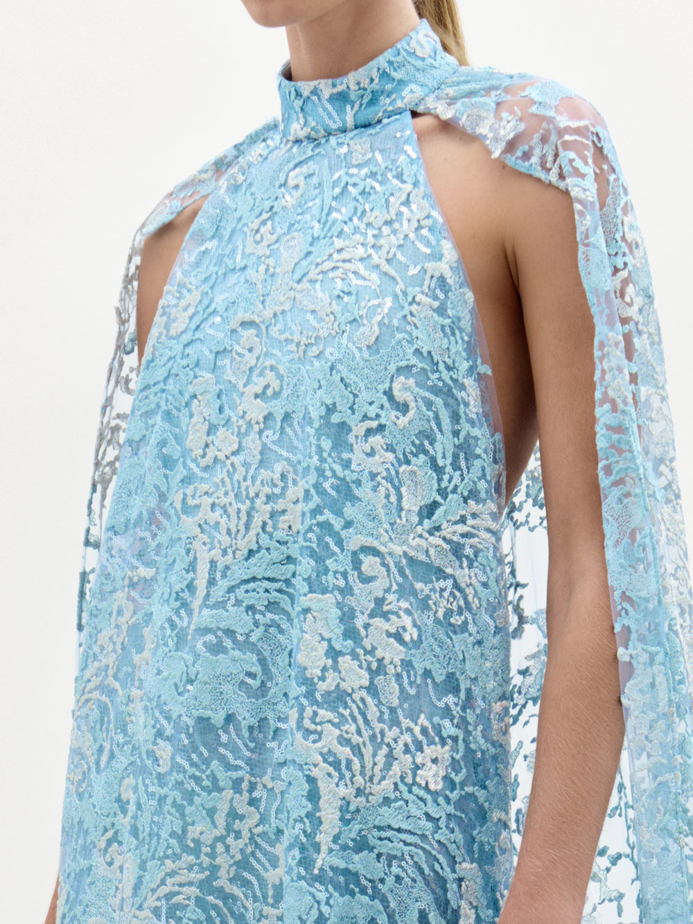 A woman models the Carol Dress—a light blue, floor-length gown made from Italian fabric, featuring a high neckline, cape shoulders, lace details, and a front slit—against a plain white background.