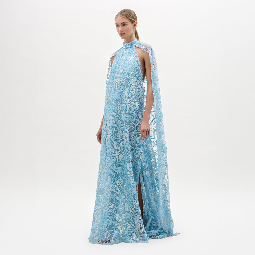 A woman models the Carol Dress—a light blue, floor-length gown made from Italian fabric, featuring a high neckline, cape shoulders, lace details, and a front slit—against a plain white background.