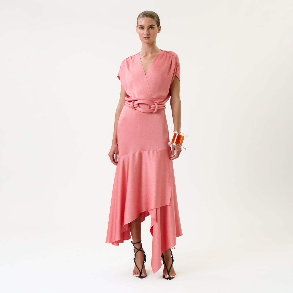 A woman wearing the Protea Dress—a pink wrap dress with an asymmetrical hem—paired with strappy black heels and an orange bracelet, stands against a plain white background.