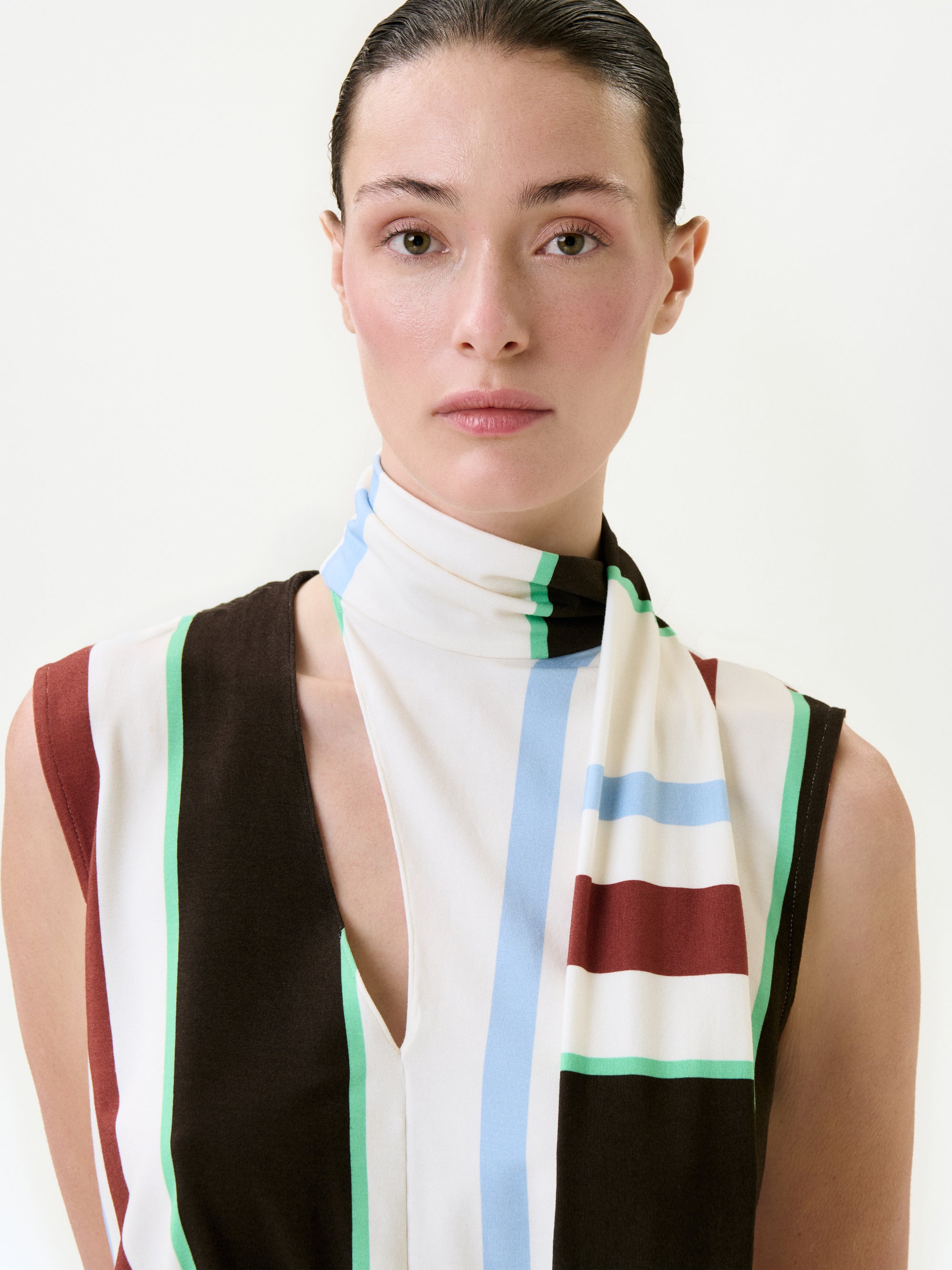 A woman with slicked-back dark hair wears the Zaz Dress, a sleeveless style crafted from Italian fabric, featuring vertical stripes in brown, white, green, blue, and red. She faces the camera with a neutral expression against a plain background.