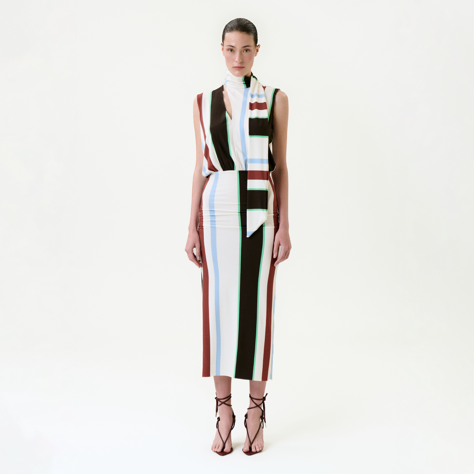 A woman stands against a white background, wearing the Zaz Dress—a sleeveless midi with vertical stripes in brown, white, light blue, and mint green. Made in Colombia from fine Italian fabric, she pairs it with a matching scarf and black heels. Her hair is pulled back.