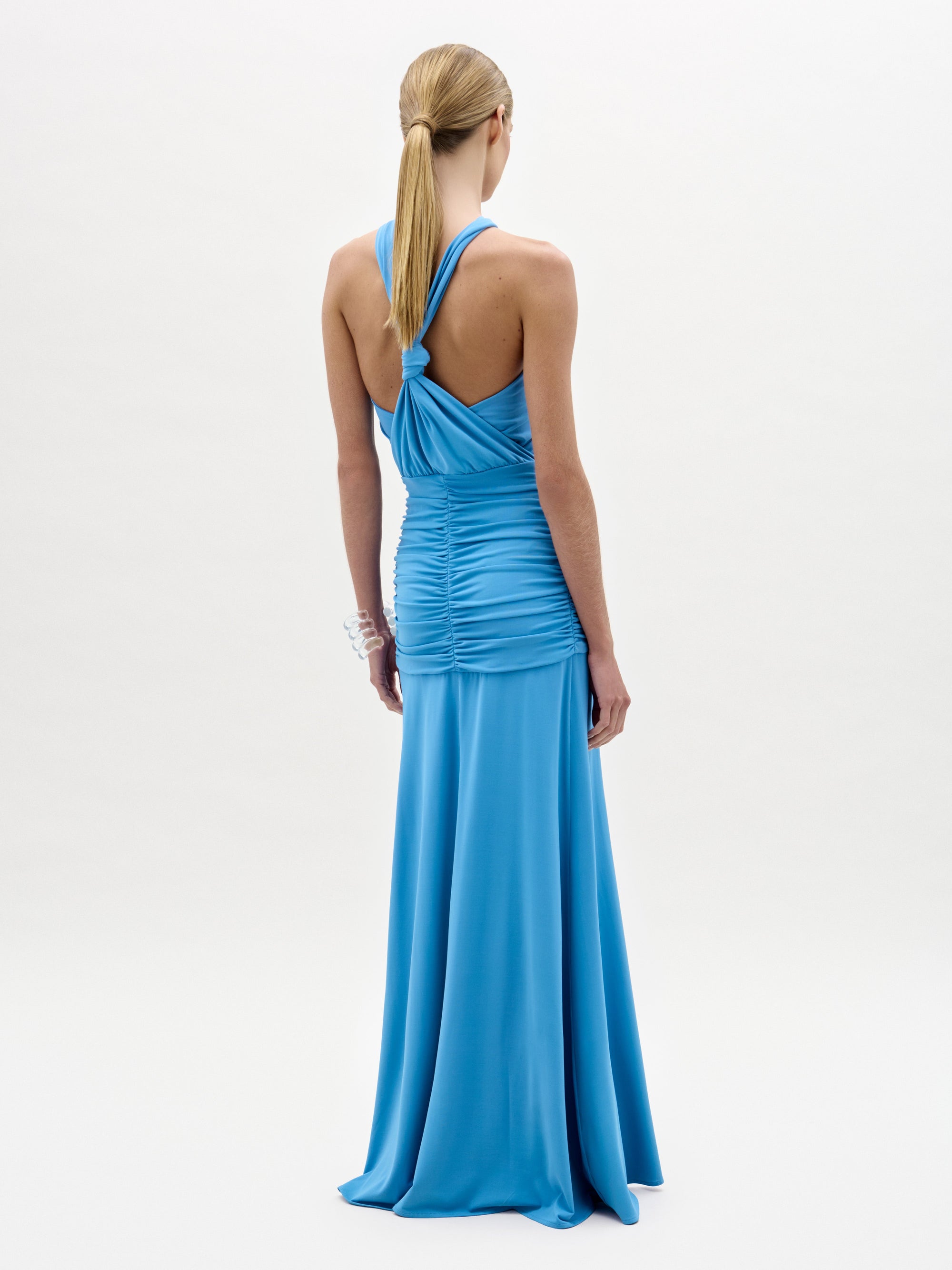 A woman wears the Jedda Dress, a sleeveless, floor-length blue maxi made from luxurious Italian fabric with ruched detailing and a fitted silhouette, paired with gold sandals against a plain white background.