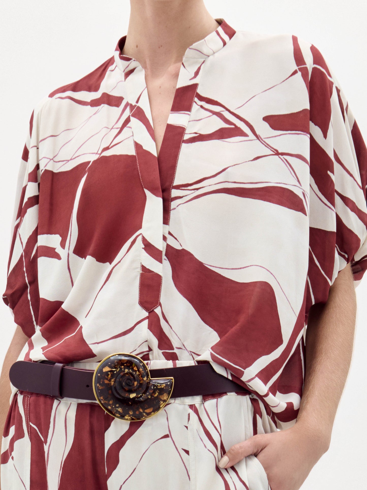 A woman poses against a plain white background wearing the Susanne Blouse, featuring a red and white abstract pattern, matched with coordinating pants and accessorized with a black belt with a gold buckle.