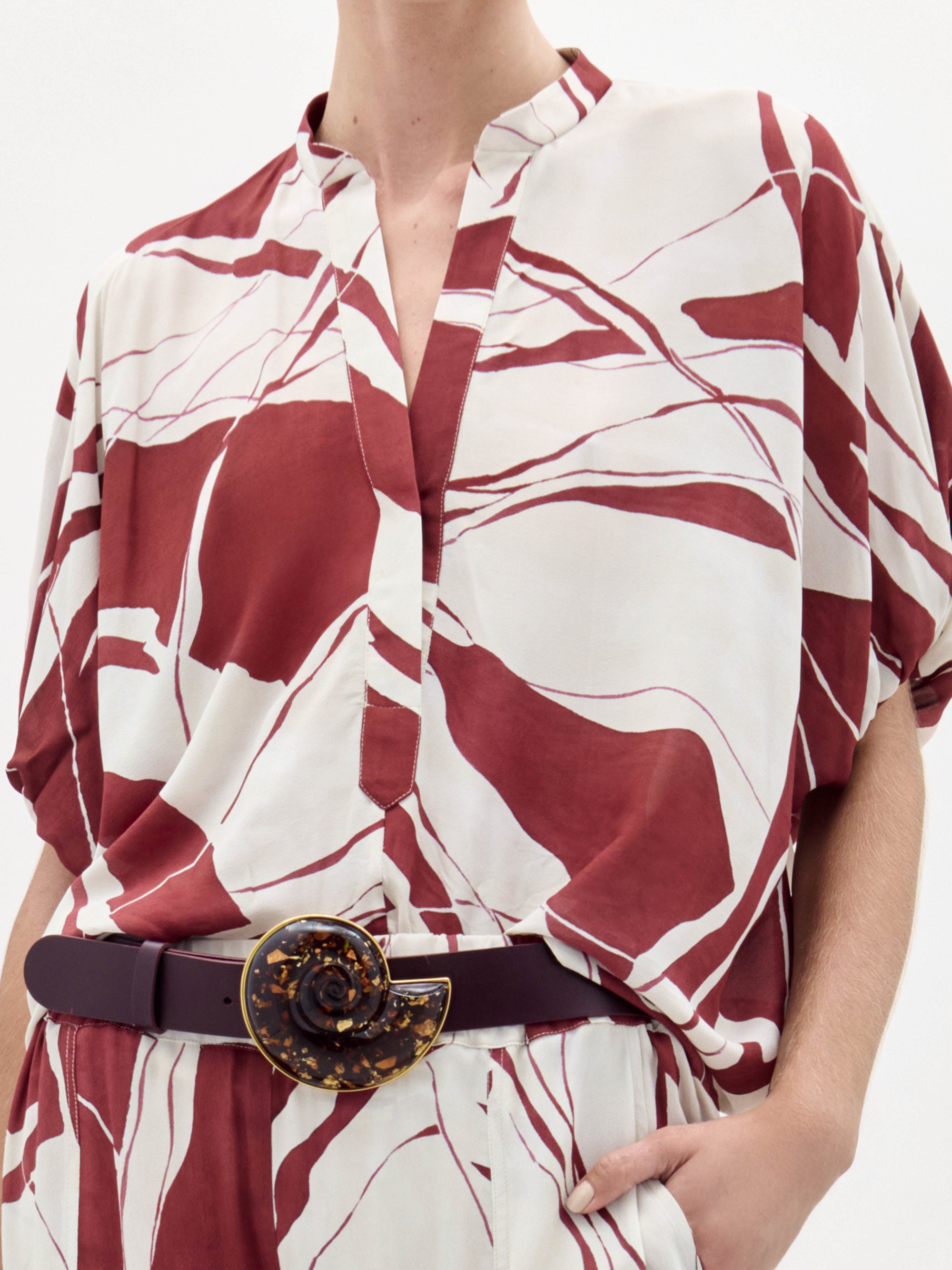A woman poses against a plain white background wearing the Susanne Blouse, featuring a red and white abstract pattern, matched with coordinating pants and accessorized with a black belt with a gold buckle.
