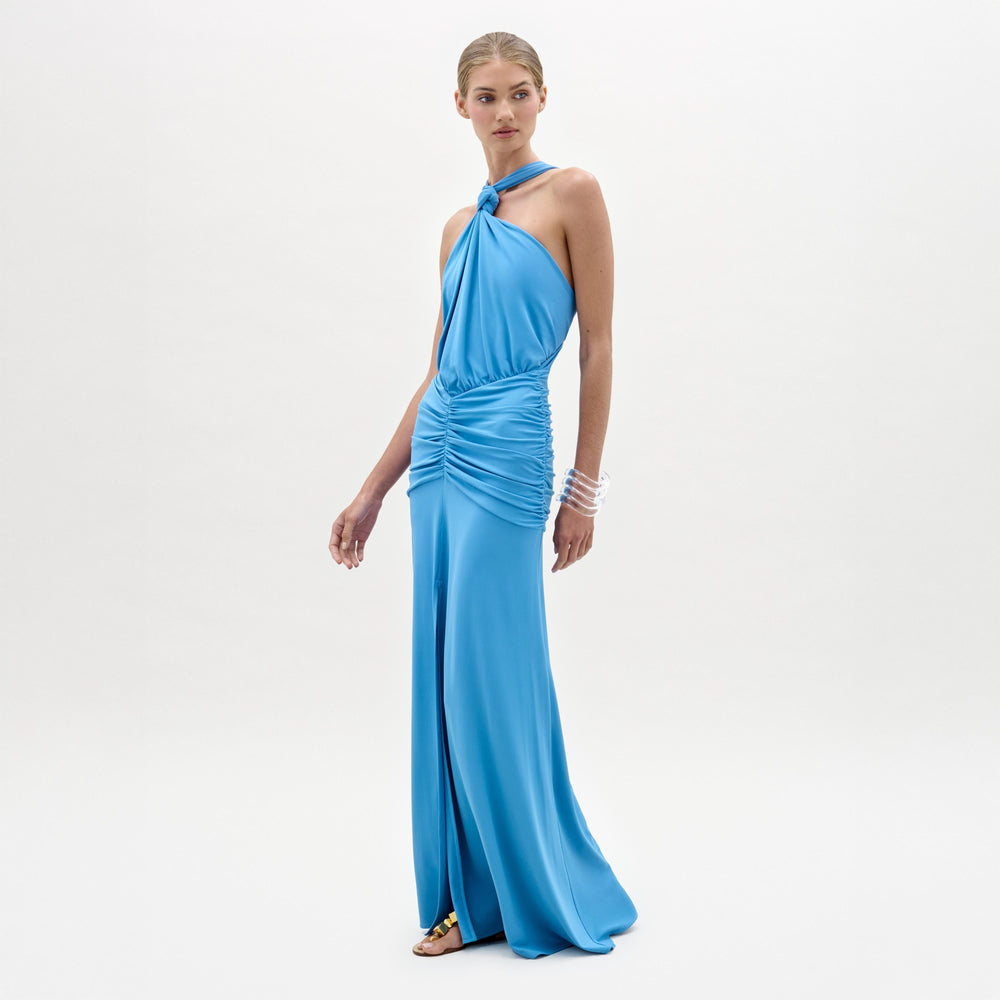 A woman wears the Jedda Dress, a sleeveless, floor-length blue maxi made from luxurious Italian fabric with ruched detailing and a fitted silhouette, paired with gold sandals against a plain white background.