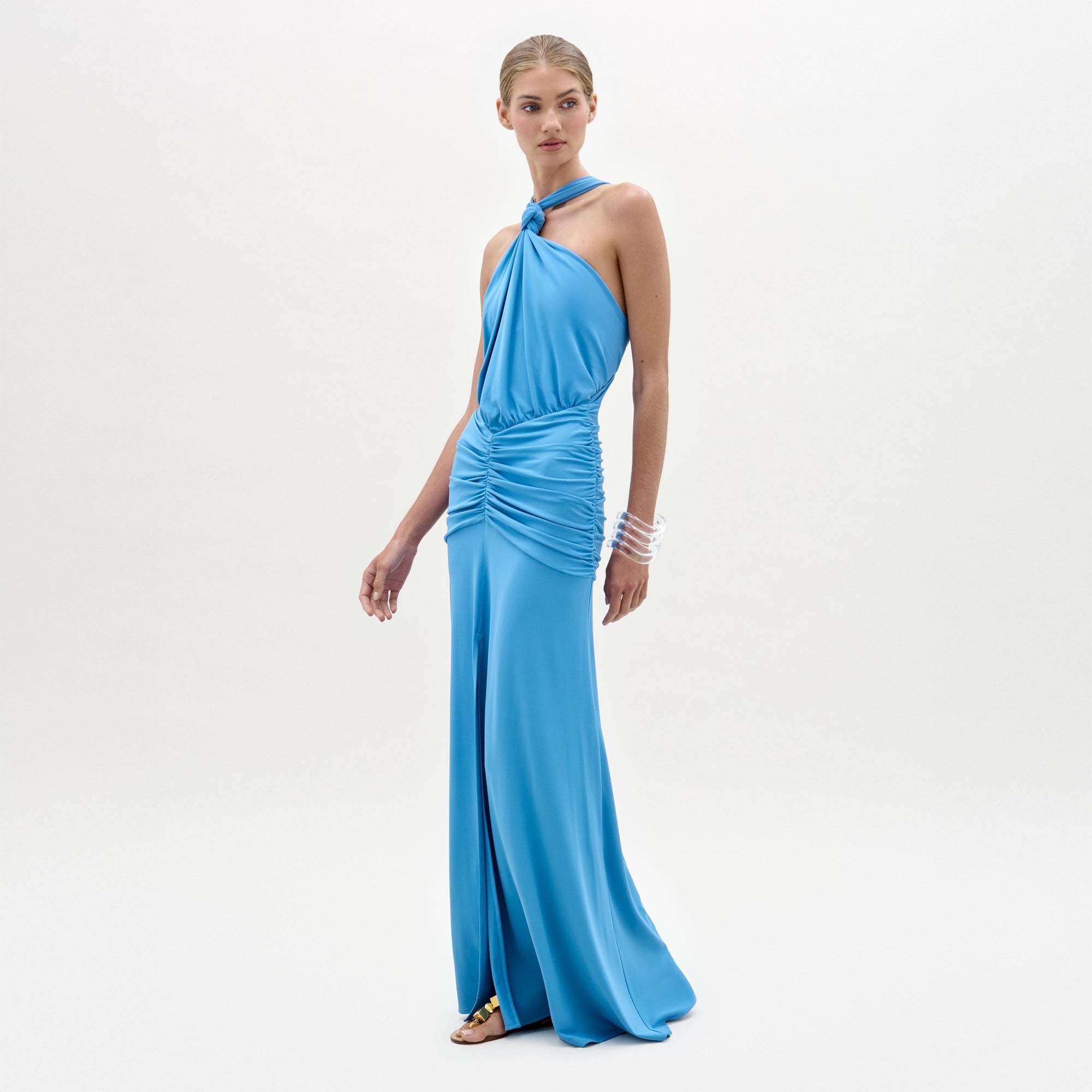 A woman wears the Jedda Dress, a sleeveless, floor-length blue maxi made from luxurious Italian fabric with ruched detailing and a fitted silhouette, paired with gold sandals against a plain white background.