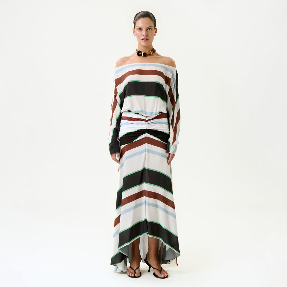 A woman wears the Sara Dress—an off-the-shoulder, long-sleeved maxi with bold brown, black, white, and green horizontal stripes—made in Colombia. She pairs it with black sandals and a chunky necklace against a white background.