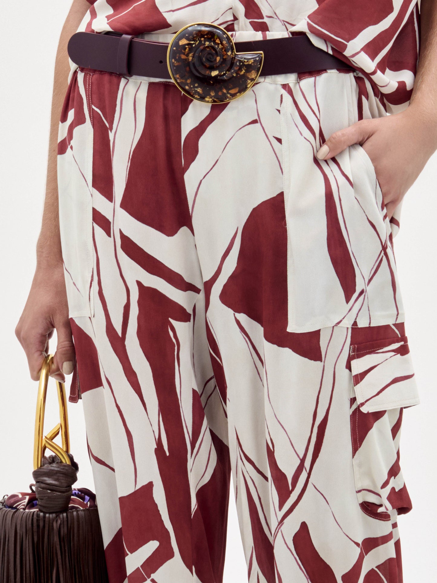 A woman stands against a plain background wearing the Jess Pant in a red and white pattern, gold sandals, a dark belt, and holding a dark fringed bag.