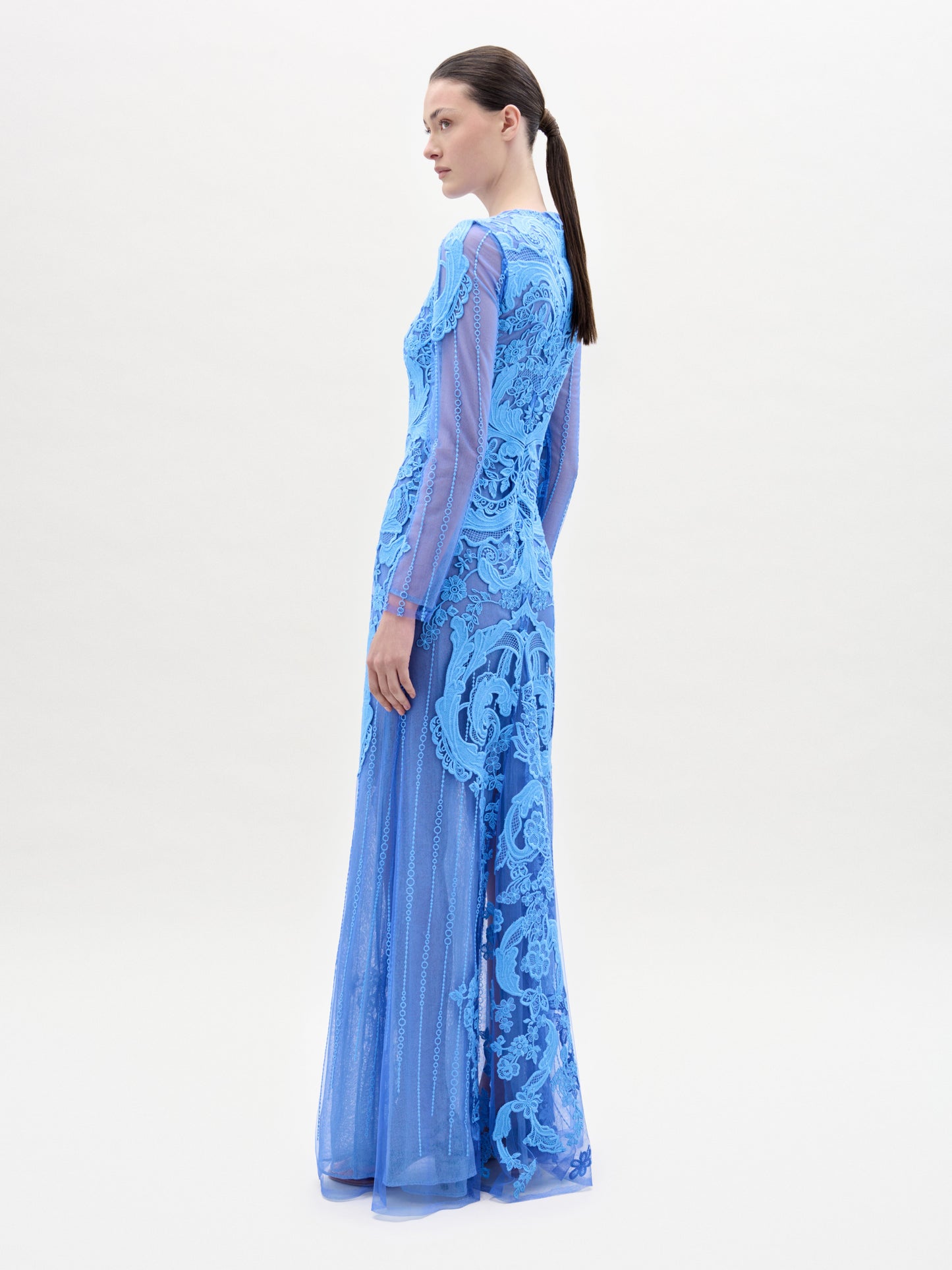 A woman stands facing forward in the Lacey Dress, a long, fitted floral lace gown with intricate patterns, long sleeves, and a beautifully tailored bodice, set against a plain light background.