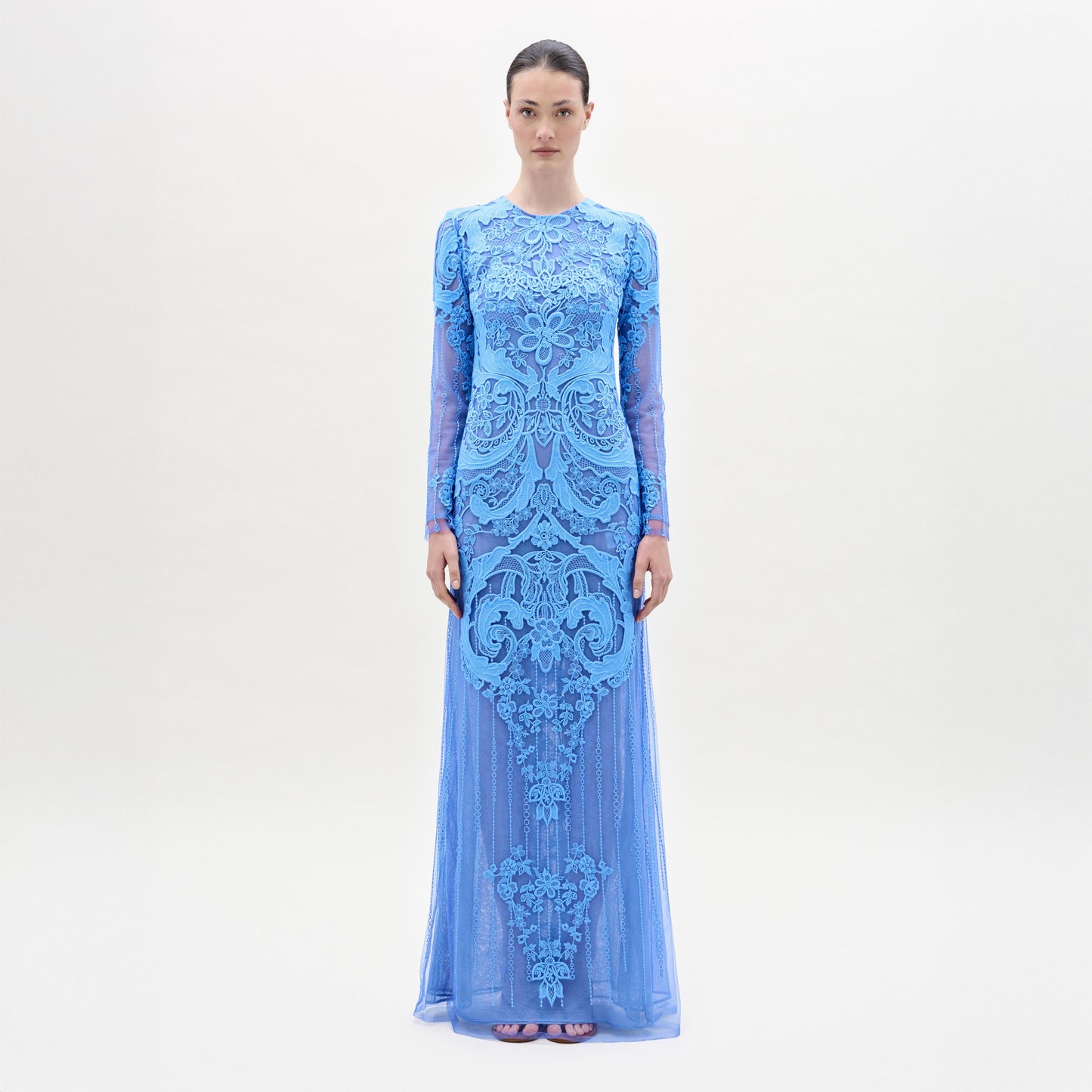 A woman stands facing forward in the Lacey Dress, a long, fitted floral lace gown with intricate patterns, long sleeves, and a beautifully tailored bodice, set against a plain light background.