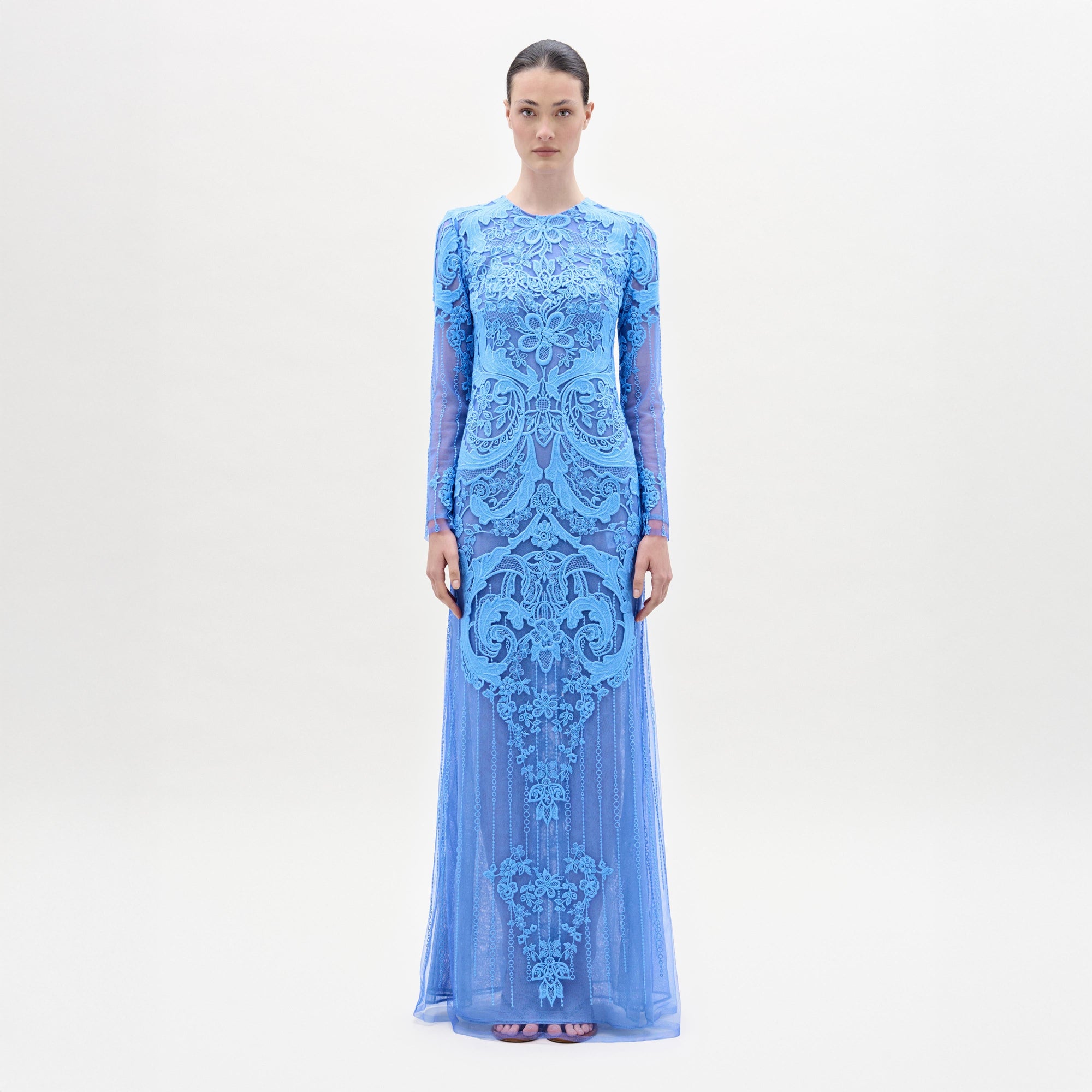 A woman stands facing forward in the Lacey Dress, a long, fitted floral lace gown with intricate patterns, long sleeves, and a beautifully tailored bodice, set against a plain light background.