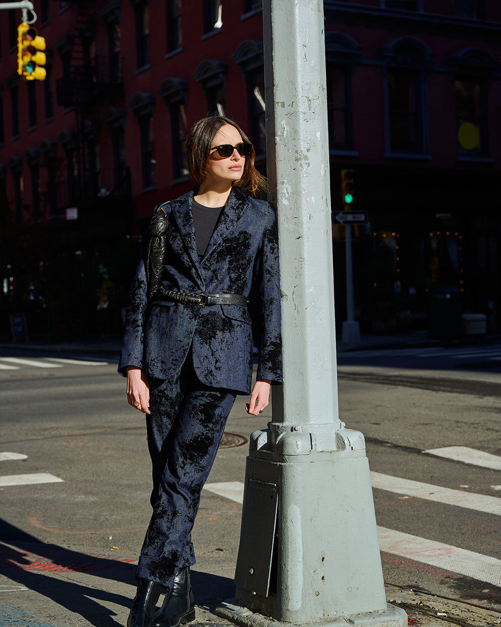 A woman wearing a dark navy crushed velvet suit with a matching belt and black boots stands on a city street next to a gray pole.
