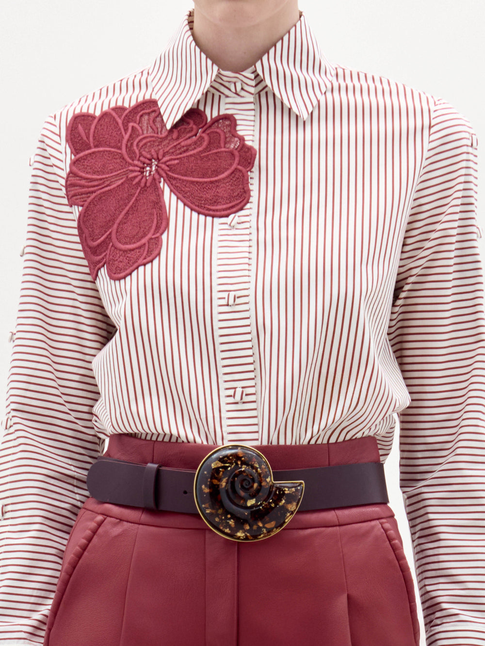 A woman stands against a plain background wearing the Premium Blouse, featuring stripes and a large red floral design, paired with red belted trousers and a decorative belt buckle.