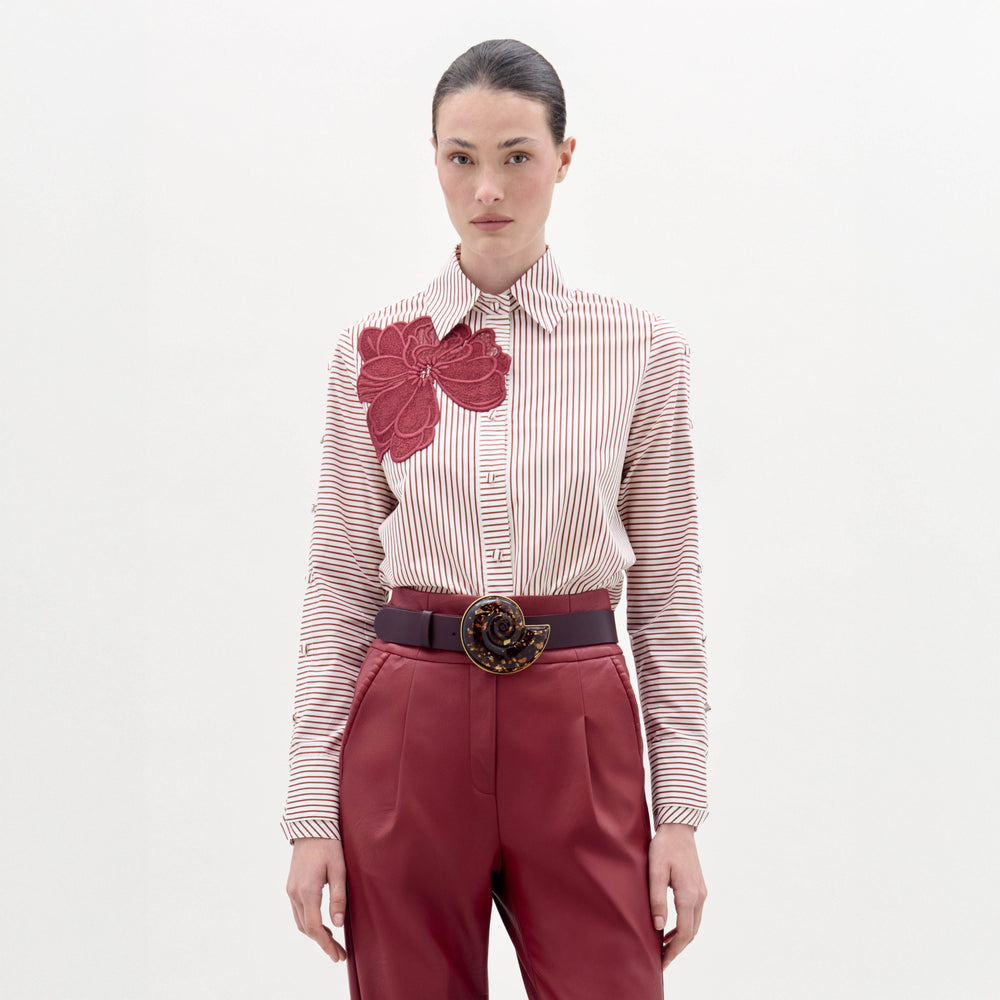 A woman stands against a plain background wearing the Premium Blouse, featuring stripes and a large red floral design, paired with red belted trousers and a decorative belt buckle.