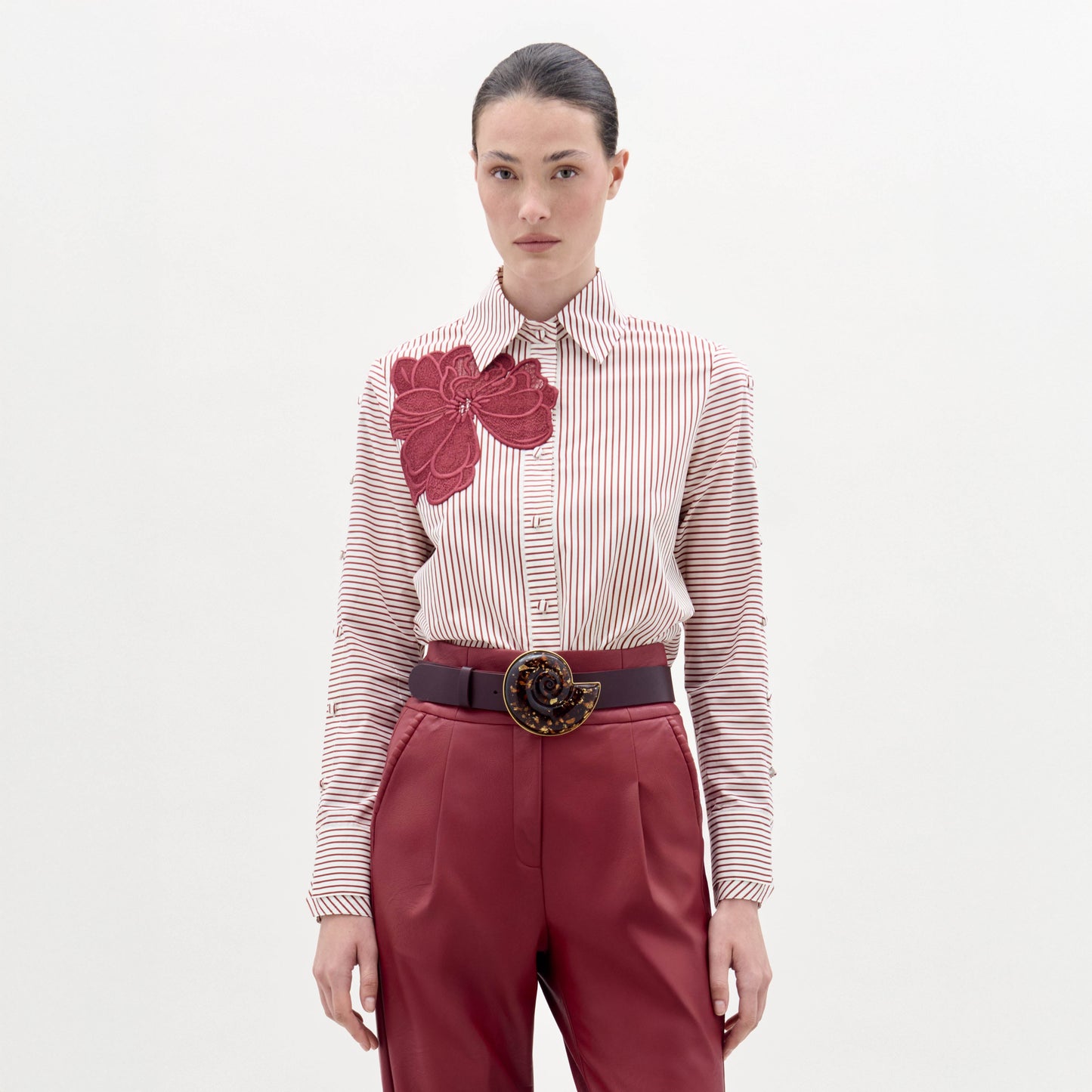 A woman stands against a plain background wearing the Premium Blouse, featuring stripes and a large red floral design, paired with red belted trousers and a decorative belt buckle.
